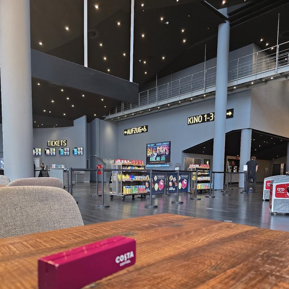 A movie theater lobby with a wooden table and a Costa Coffee box. There are posters for